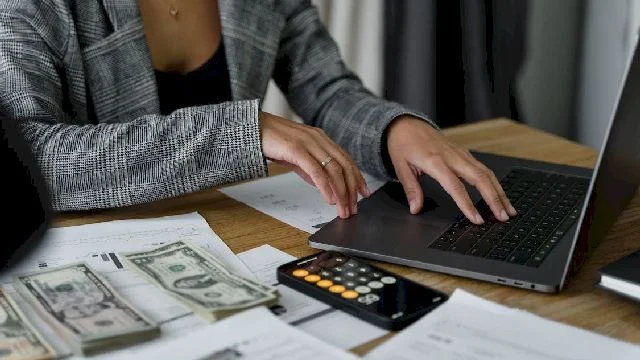 Image of a woman working on a laptop on a table with cash notes and papers.